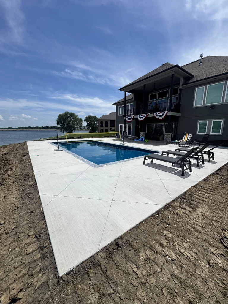 Rectangular fiberglass pool with wide concrete deck behind a dark home