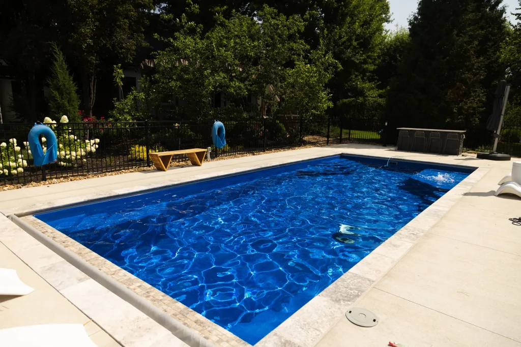 Rectangular fiberglass pool with travertine coping in a wooded backyard